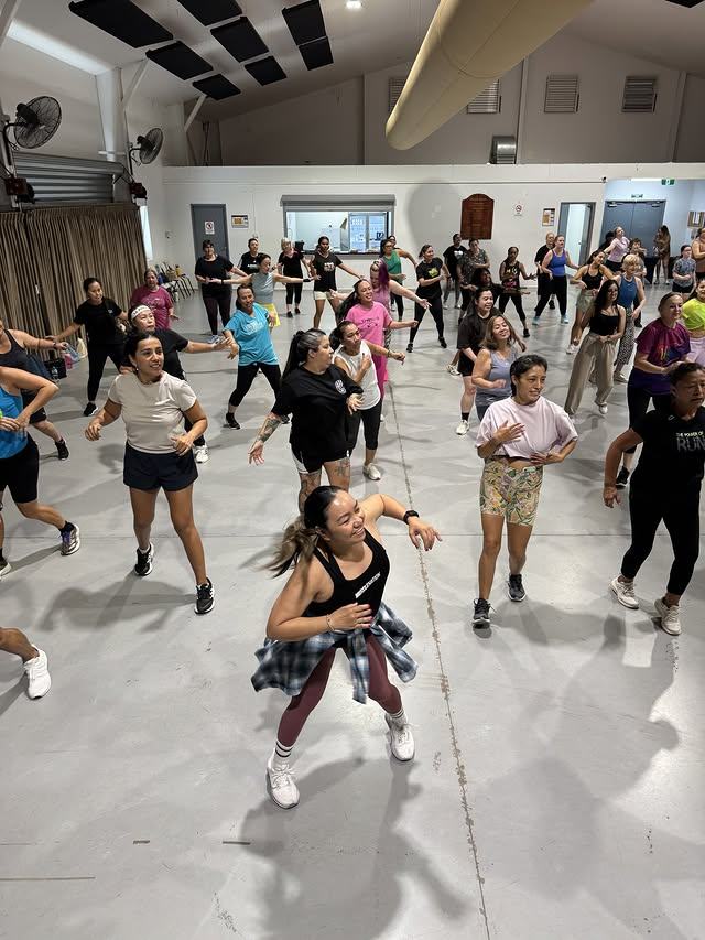 A full class mid-routine in a community hall, arms raised together.